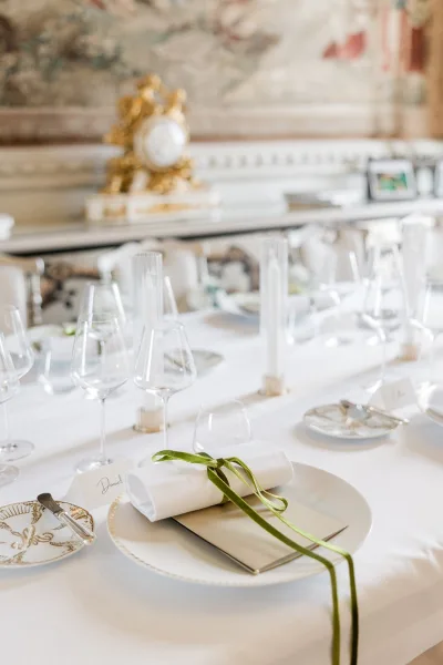 Wedding place setting with a green ribbon napkin and menu card, set against a tapestry wall with a gilded clock and mantel behind.