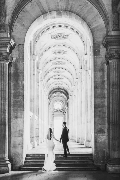 Couple portrait of the bride and groom holding hands under stone archways and columns in a grand hallway, her veil trailing on the steps.