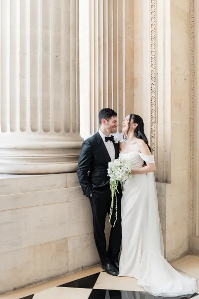 Couple portrait of a bride and groom laughing, she holds a white and green bouquet as they stand by stone columns and a checkered floor.
