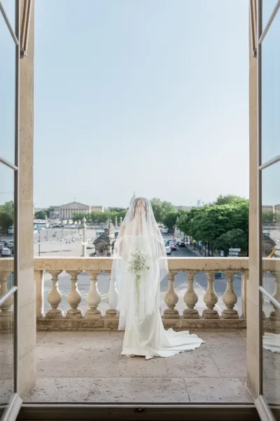 Bridal portrait of a bride on a stone balcony, framed by open doors, her flowing veil catching light against a historic cityscape and blue sky.