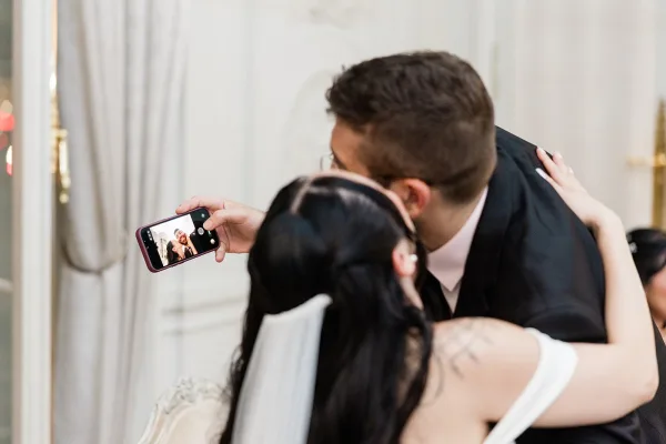 Wedding selfie of the bride and groom as they share a kiss, the bride holding a smartphone close while he leans in.