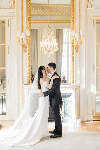 Wedding kiss portrait as the bride and groom kiss beneath a chandelier, framed by tall windows and paneled walls in an elegant room.