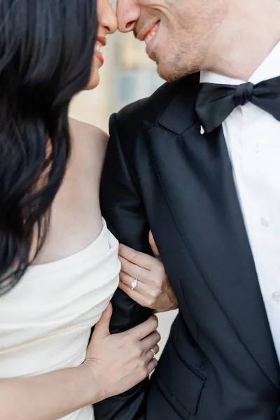 Couple portrait of a bride holding the groom’s arm as they nearly kiss by a stone wall, her diamond engagement ring catching light.