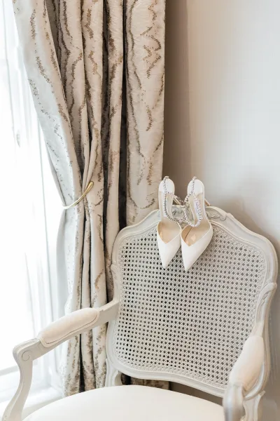 Bridal shoes, ivory wedding heels with rhinestone ankle straps, arranged against patterned drapes, bathed in soft window light.