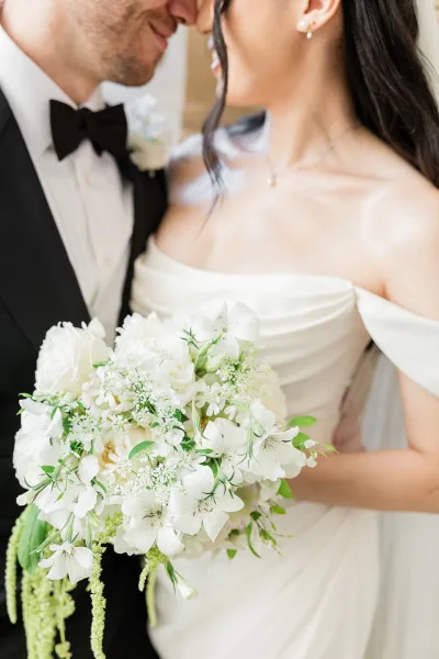 Couple portrait of a bride holding a bouquet, noses almost touching her groom in a black tuxedo, white and green florals trailing elegantly.