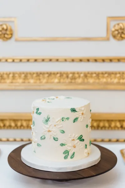 Wedding cake, a single-tier design with buttercream florals, displayed against a white wall with gold molding on a clothed table.
