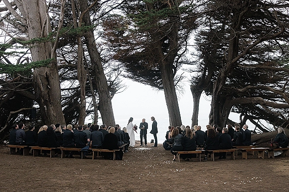 Outdoor wedding ceremony with bride and groom before an officiant, wood bench seating lining a petal-strewn aisle, ocean view beyond trees
