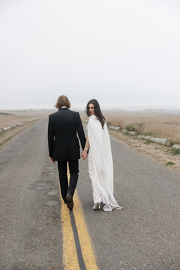 Couple portrait of bride and groom walking away holding hands on an empty coastal road, bride looking back in cape veil under overcast sky