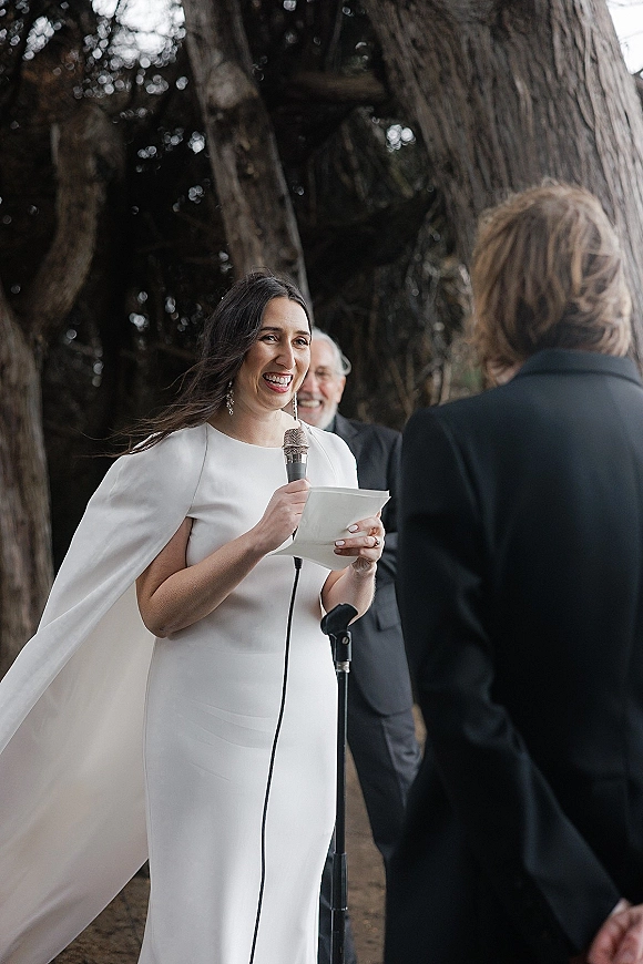 Wedding vows as the bride reading vows into a handheld microphone, holding vow papers in a cape-sleeve white gown under trees outdoors
