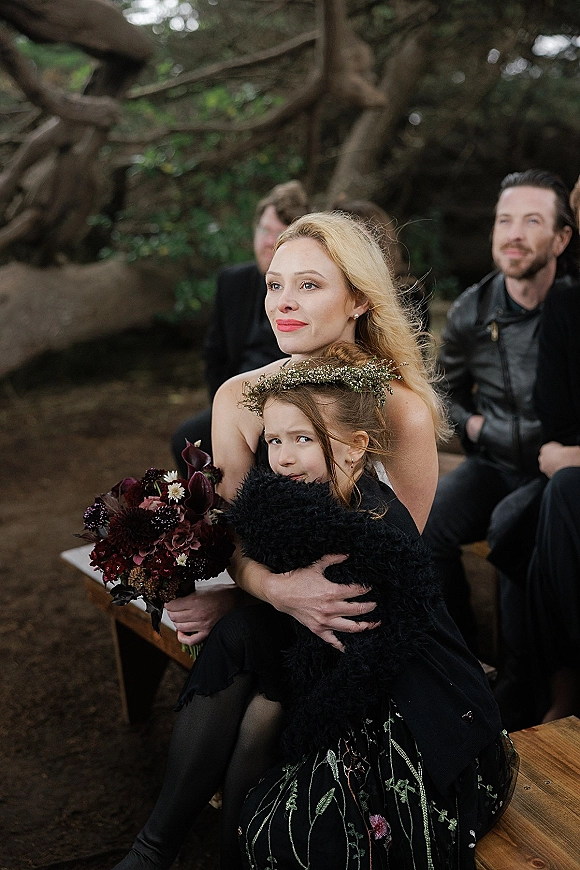 Wedding guest portrait of a woman with a dark floral bouquet and flower crown on a wooden bench under tree branches as guests sit behind