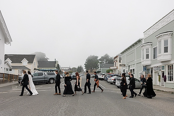 Wedding party crossing street, bride and groom walking with a long veil as bridesmaids in black and groomsmen in dark suits pass storefronts in foggy overcast light