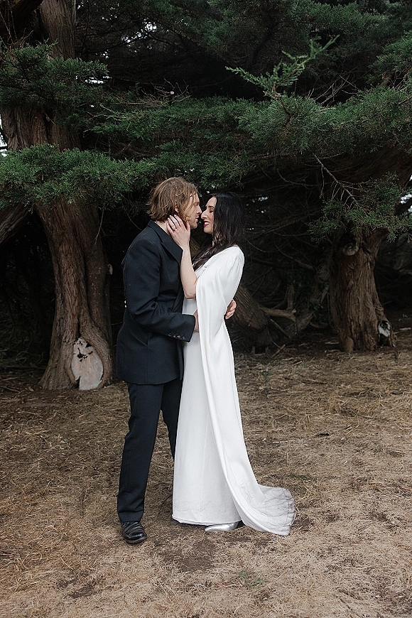 Couple portrait of bride and groom embracing as she touches his face, wedding dress and suit framed by evergreen trees and grass