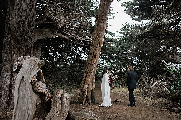 Couple portrait of bride and groom facing each other on a forest path under tall trees, holding a dark red bouquet in moody light