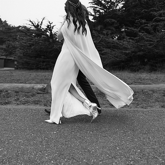 Wedding couple portrait in black and white of bride and groom walking from behind, bridal cape flowing along a tree-lined path by a log fence