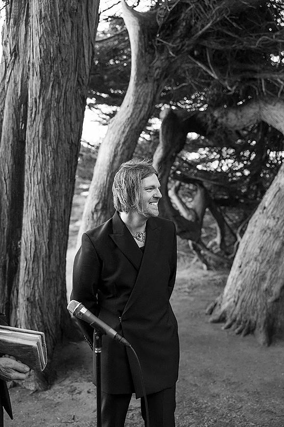 Groom portrait in a black suit and necklace, smiling by a microphone on a forest path at an outdoor wedding ceremony