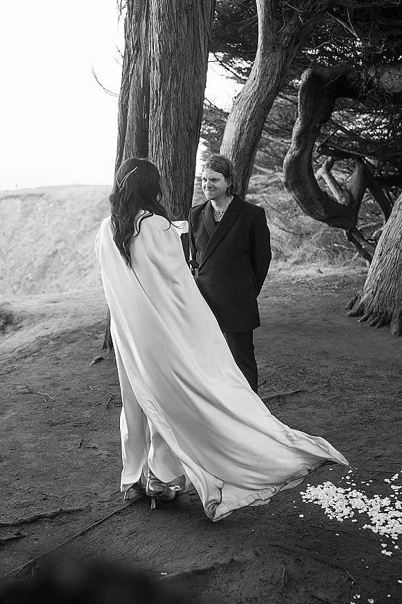 First look moment as bride in a windswept bridal cape approaches groom on a dirt path under trees with coastline and rose petals