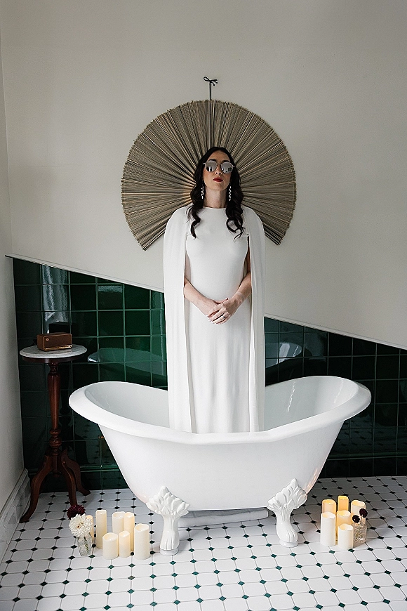 Bridal portrait of a bride in sunglasses wearing a cape-sleeve wedding dress by a clawfoot tub with candle and flower vases, green tile wall backdrop