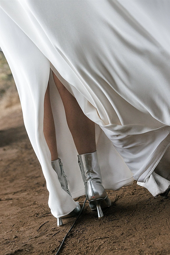 Bridal shoes in silver wedding boots peek from a white satin dress hem, showing high heels and zipper detail on dirt ground