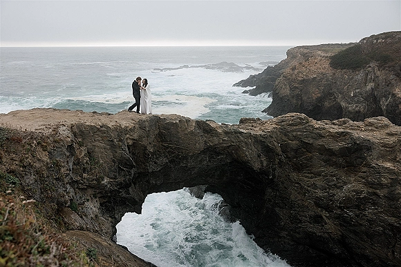 Couple portrait of bride in a flowing veil and groom in suit standing on a rocky cliffside with ocean waves and a sea arch under overcast sky