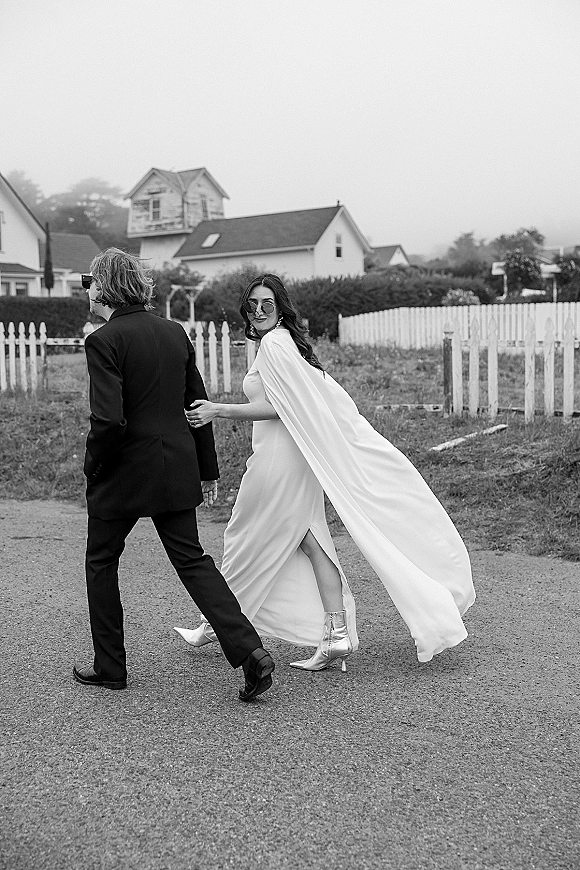 Couple portrait of bride and groom walking on a gravel road, bride looking back in cape veil, sunglasses, and ankle boots under overcast sky