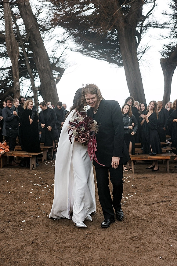 Wedding recessional as bride and groom walk the aisle, bouquet in hand, flower petals falling amid cheering guests under trees and overcast sky