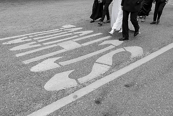 Wedding processional with bride and groom walking across a city crosswalk, bridal gown hem and heels visible as guests follow behind