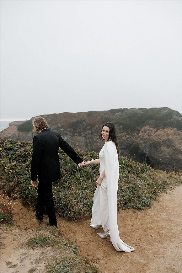 Couple portrait of a wedding couple holding hands on coastal cliffs, bride looking back in a white cape-sleeve gown with train, groom in black suit