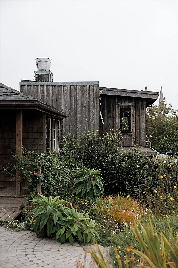 Rustic barn exterior with weathered wood siding, window, and porch posts beside wildflowers and a stone paver path under an overcast sky
