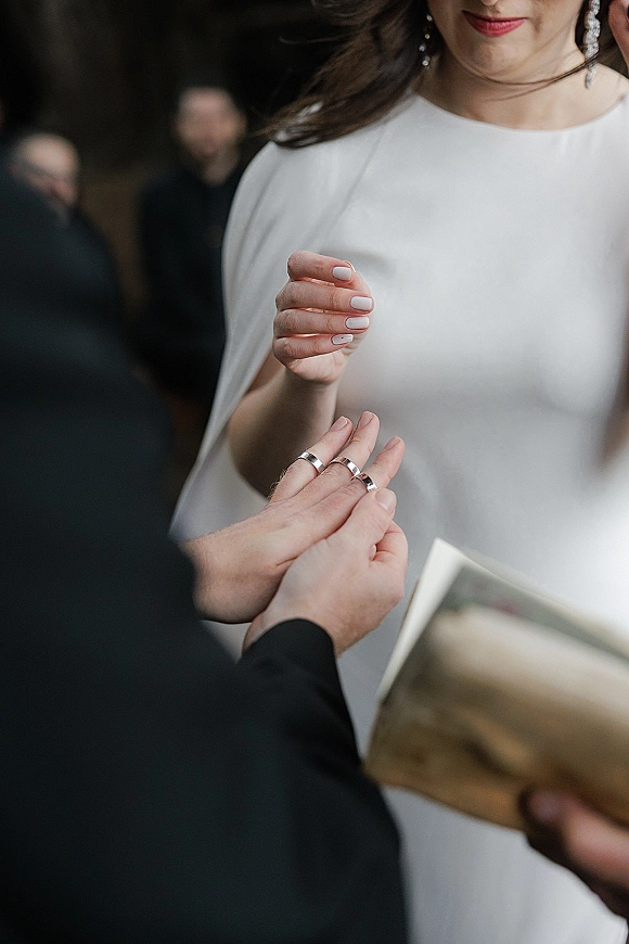 Wedding ring exchange as the bride slides a band onto the groom’s hand, light pink manicure and drop earrings, officiant holding a book behind
