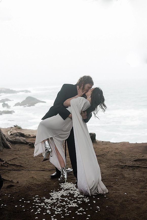 Wedding kiss portrait with groom dipping bride in cape-sleeve dress, black suit, and silver boots on a rocky cliffside above the ocean