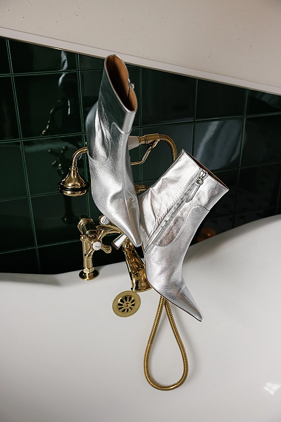 Wedding shoes—silver bridal boots with zipper detail resting on a white bathtub beside a gold faucet and handheld shower, green tile wall behind