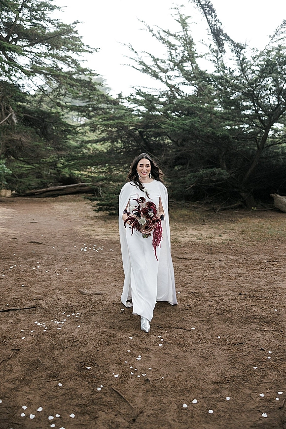 Bridal portrait of a bride walking with bouquet, wearing a white cape wedding dress and metallic ankle boots on a forest dirt path with petals