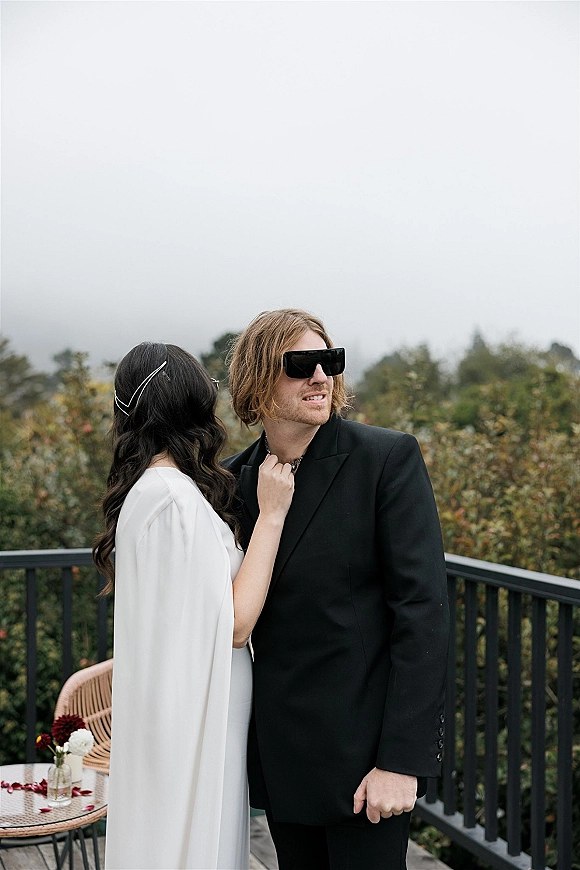 Couple portrait of bride in white cape dress adjusting groom’s collar as he wears statement sunglasses on an overcast deck with trees behind