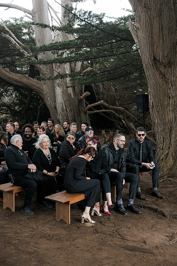 Wedding guests seated on wood benches in black attire, some wearing sunglasses, laughing during a forest ceremony under large trees