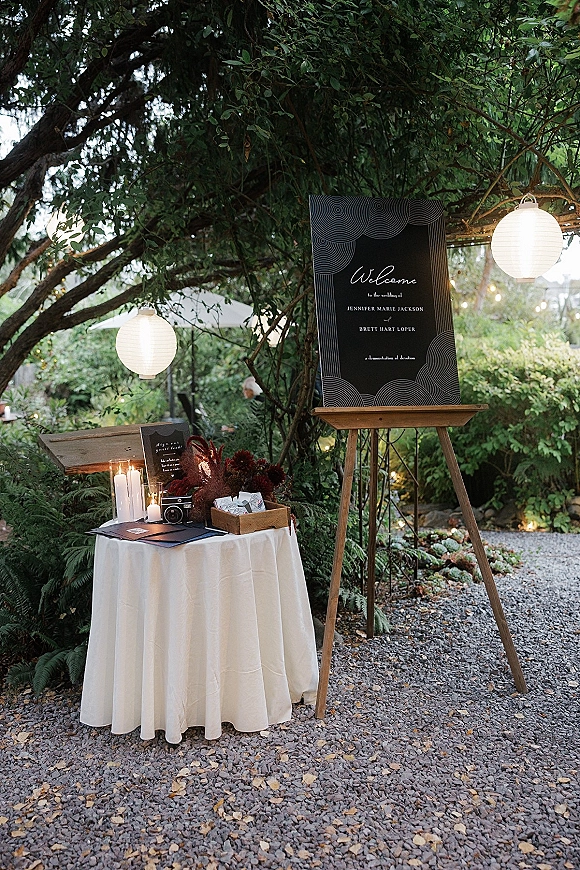 Wedding welcome sign on an easel beside a candlelit table with florals, place cards, and vintage camera under string lights in a garden setting