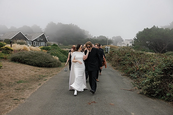 Wedding processional with bride and groom walking on a foggy paved path, bride in cape veil and sunglasses, groom in black suit with guests behind