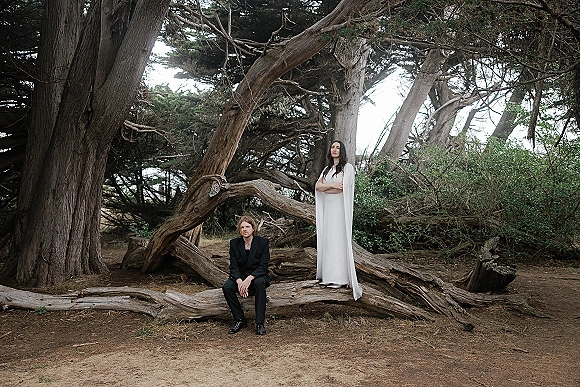 Couple portrait in a forest wedding portrait, bride in a cape sleeve dress standing beside groom in a black suit seated on fallen branches near a huge tree