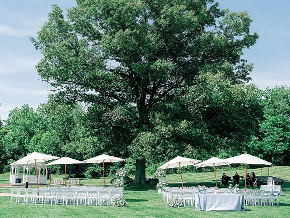 Outdoor ceremony setup with garden wedding ceremony chairs in a semicircle, white umbrellas, aisle florals, and a gazebo under a large tree