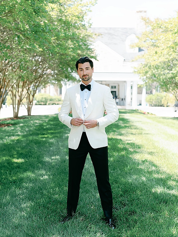 Groom portrait in a white tuxedo jacket with black bow tie, standing on a sunlit lawn with leafy trees and a white building behind