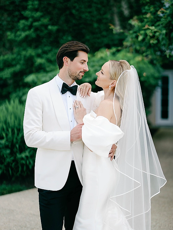 Couple portrait of bride and groom pose as she holds his lapel, off-shoulder gown and veil beside greenery on a garden path