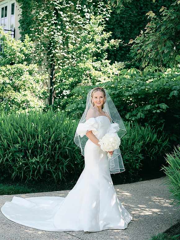 Bridal portrait of a bride holding a white hydrangea bouquet in an off-the-shoulder gown with cathedral veil along a garden path