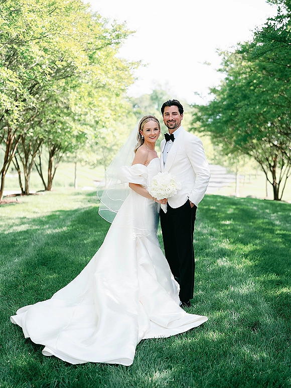 Couple portrait of bride and groom smiling, bride in veil holding white bouquet beside groom in white tuxedo on a tree-lined lawn