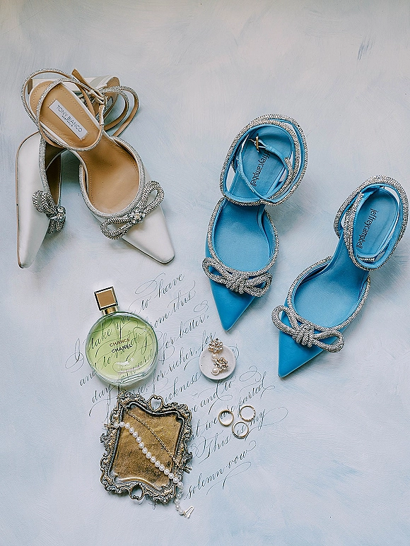 Wedding flat lay of bride getting ready details with blue heels, rings, earrings, perfume and pearl bracelet on white fabric with calligraphy paper