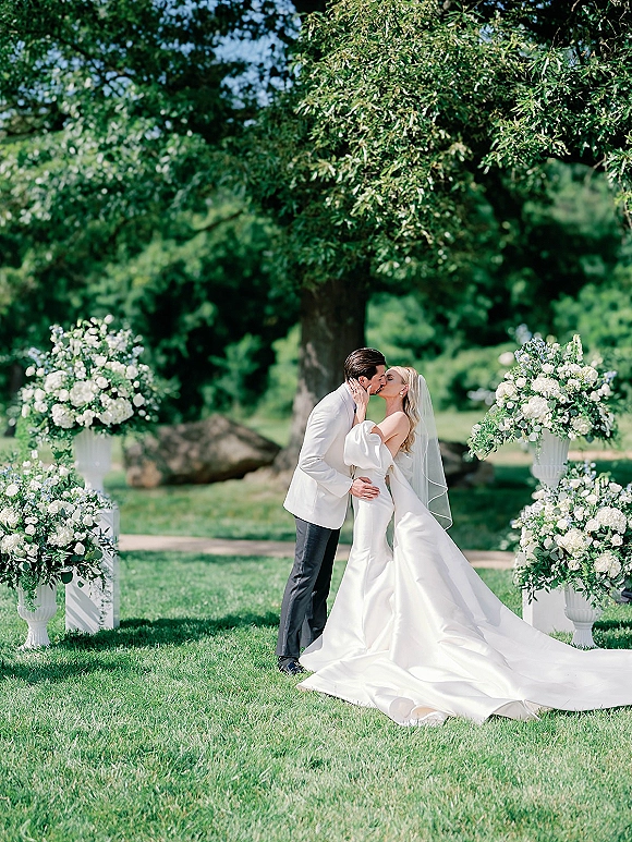 Wedding kiss portrait of bride and groom kissing, her veil flowing as they embrace on a garden lawn by white urn florals and trees