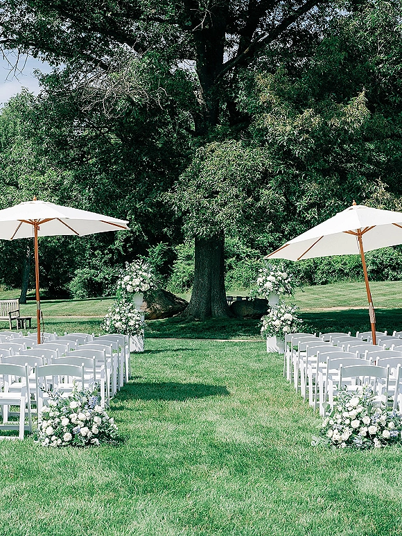 Ceremony setup for an outdoor wedding ceremony with white folding chairs, white rose and greenery aisle florals, and pillars beneath a large tree