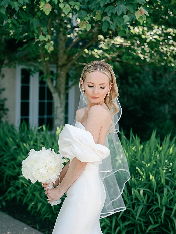 Bridal portrait of a bride in an off the shoulder wedding dress with cathedral veil and white rose bouquet on a garden walkway near a house exterior