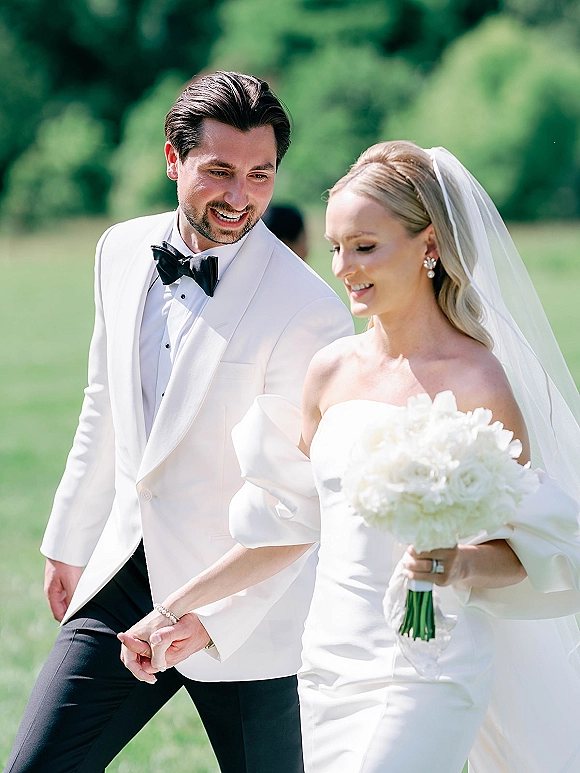 Couple portrait of bride and groom walking hand in hand, bride holding a white rose bouquet, on a green lawn with trees behind them