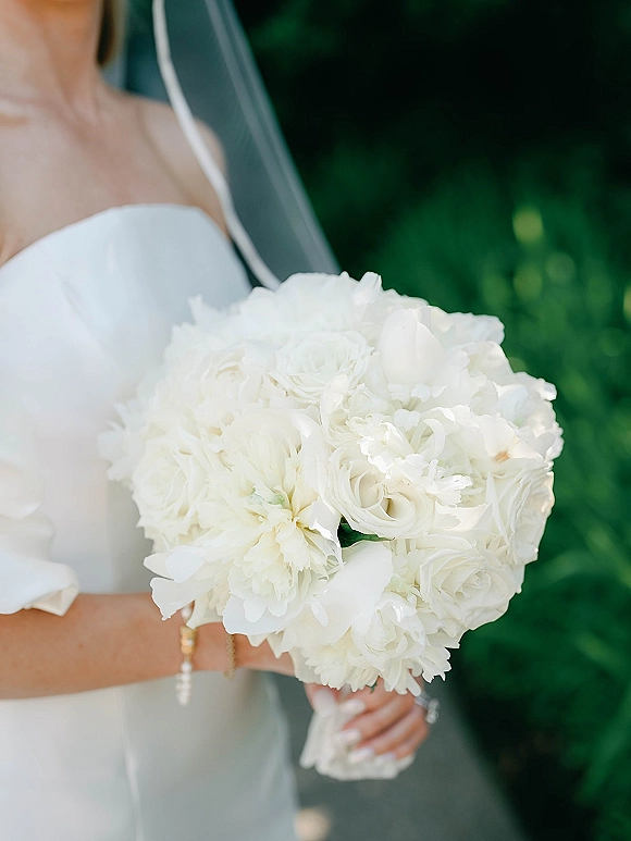 Bridal bouquet of white peonies and roses held by a bride in a strapless wedding dress and veil, with green foliage behind