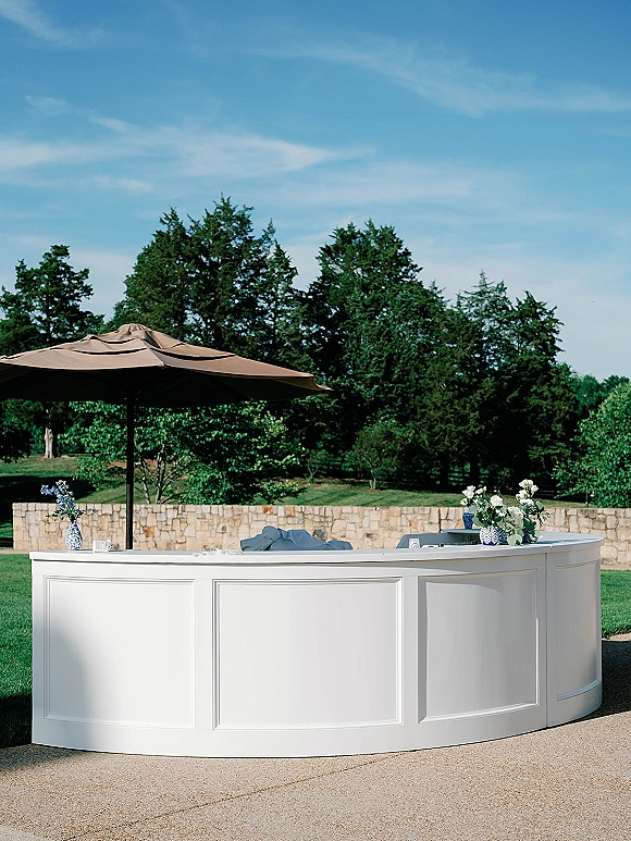 Outdoor bar setup with a white bar counter under a patio umbrella, styled with blue and white vases and white florals on a sunny lawn