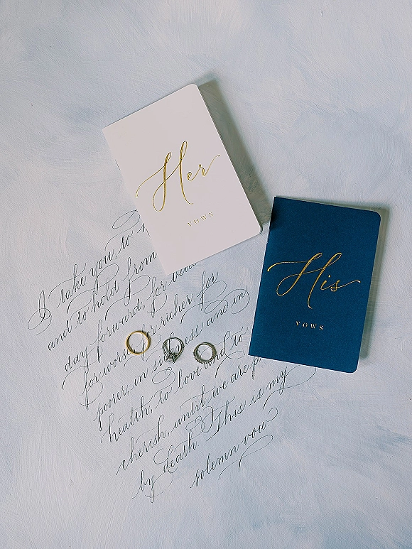 Vow books with his and her vow books in navy and white, gold foil lettering, handwritten vows, and rings on a light blue surface